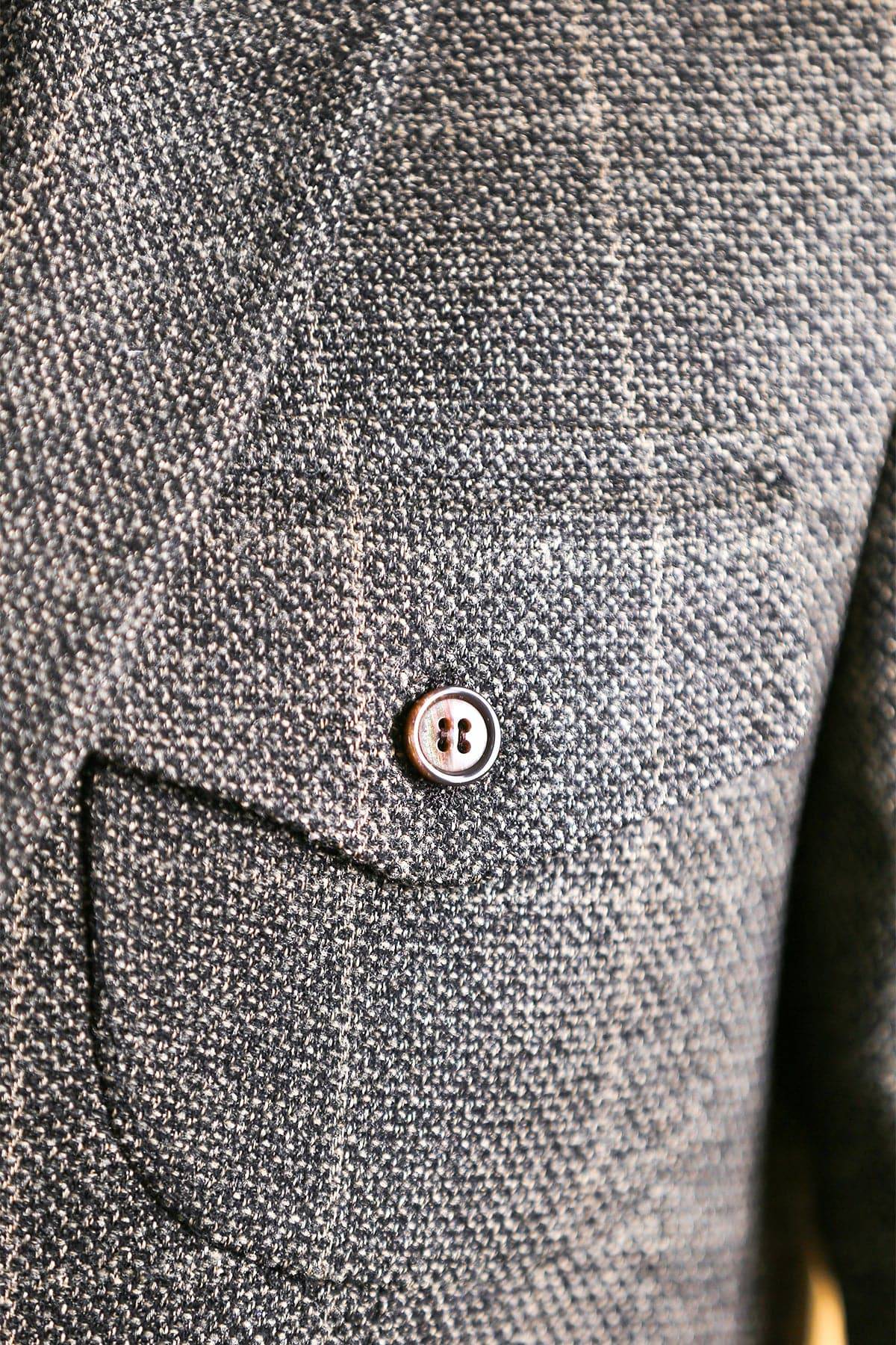 Men's blazer pocket flap detail in dark gray textured fabric with metal button.