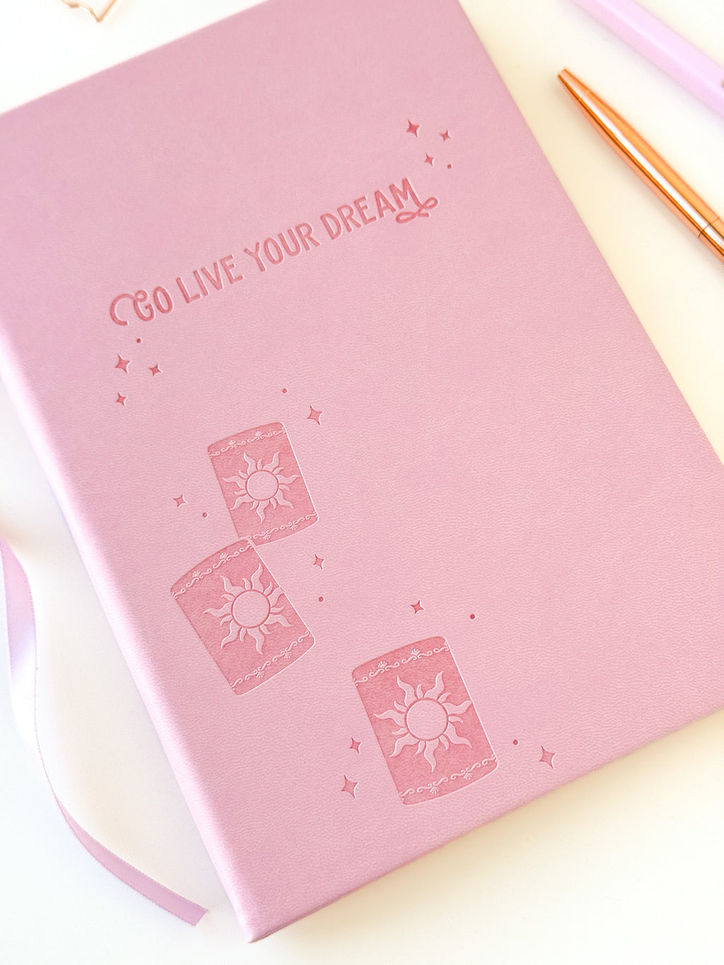 Pink notebook cover with embossed sparkle and sun icon design, lavender ribbon