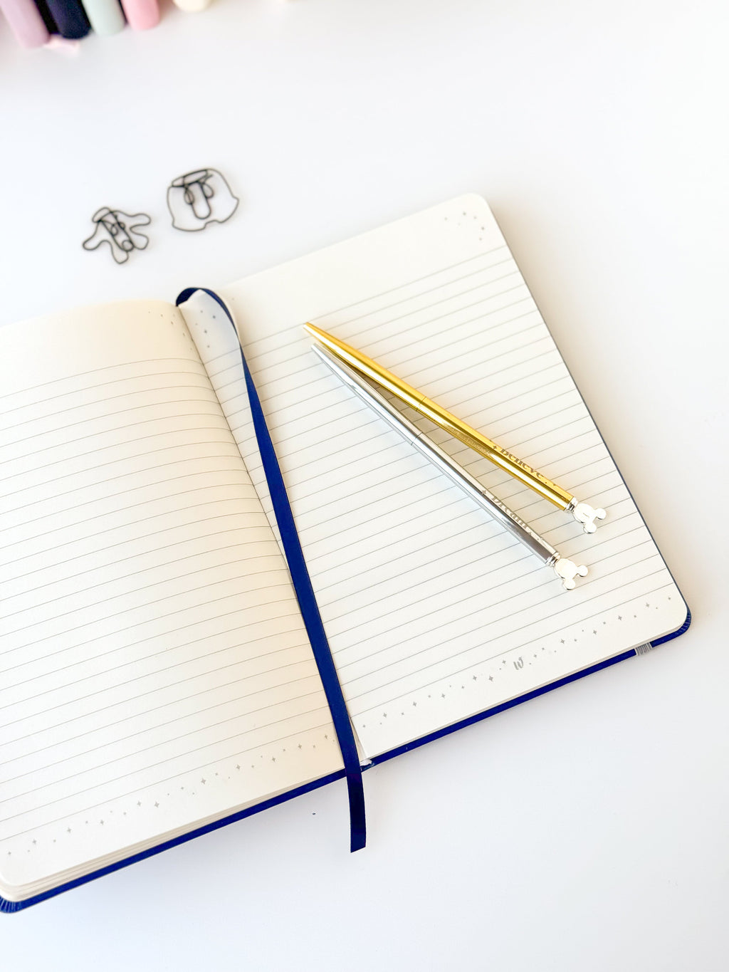 Open blue notebook with white ruled pages, ribbon spine, and gold pens