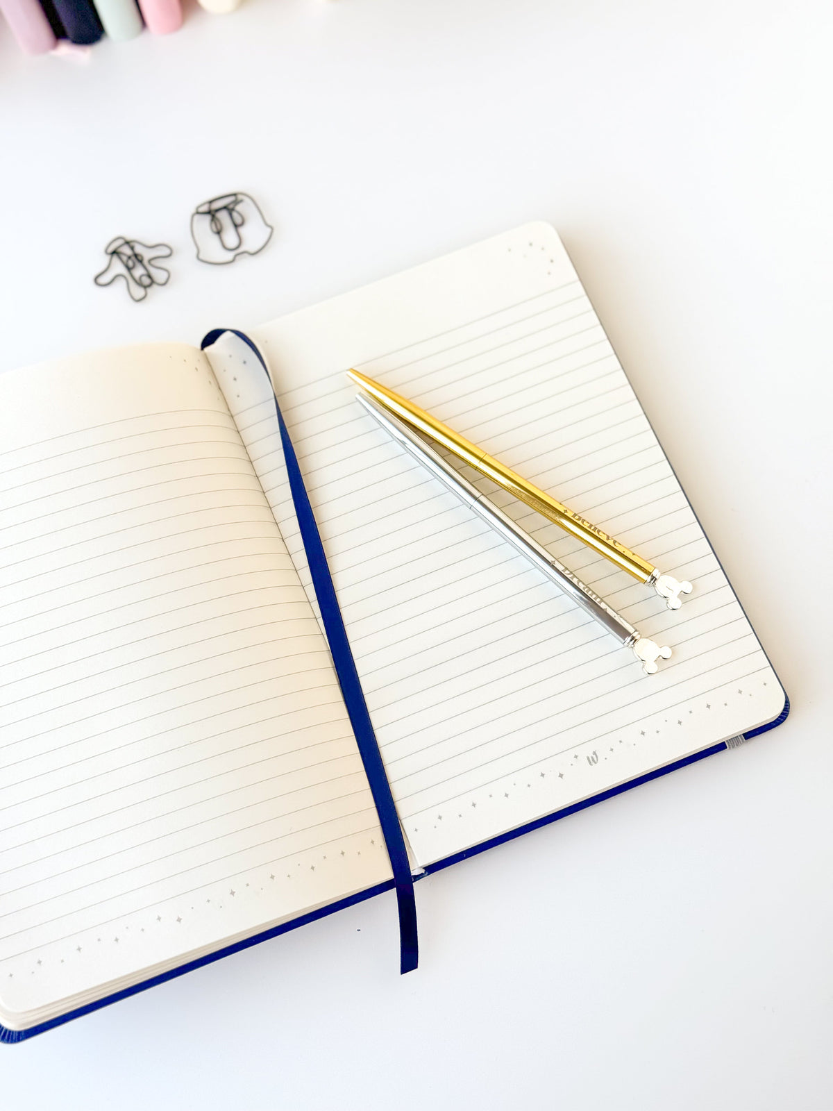 Open blue notebook with white ruled pages, ribbon spine, and gold pens
