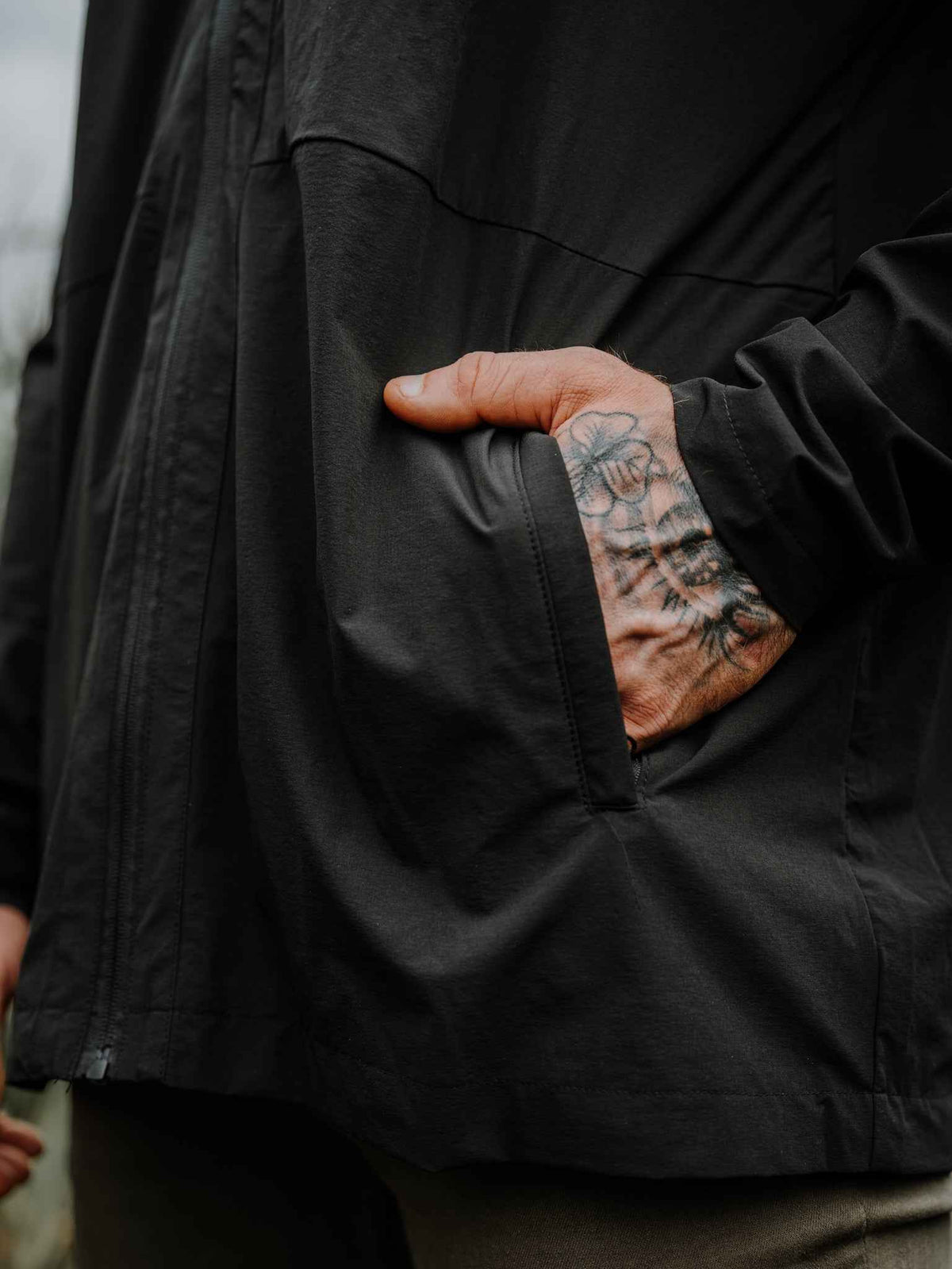 Black jacket pocket detail with stitched seam and visible tattoo on finger