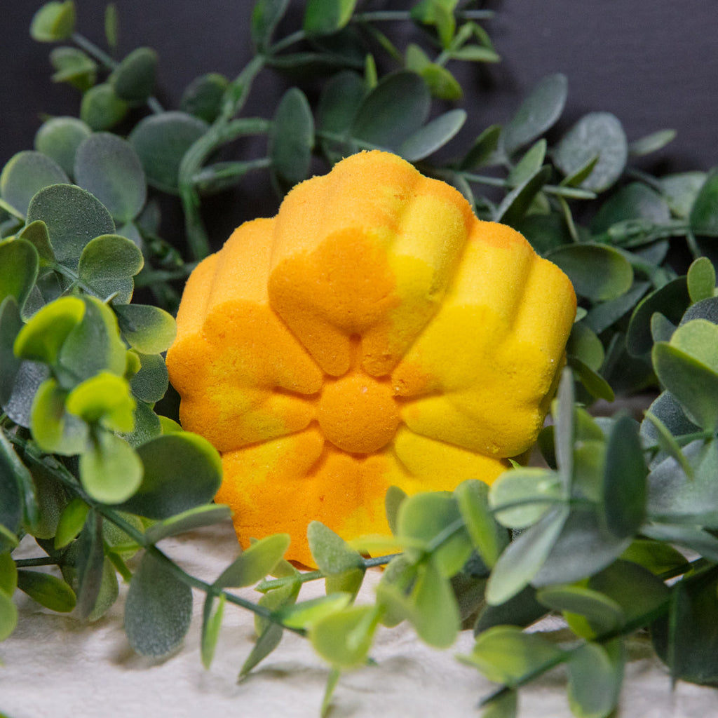 Yellow flower-shaped bath bomb in a bed of green leaves and white filler