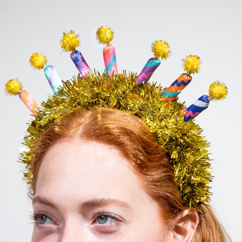 Gold tinsel headband with rainbow candle cutouts and yellow pom-pom topper details