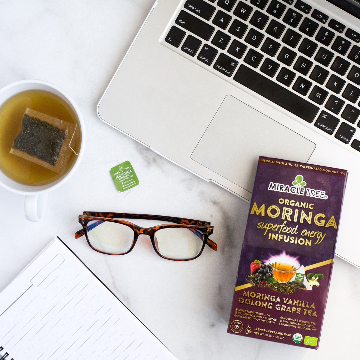 Desk setup with glass cup of tea, pyramid sachet, and Miracle Tree moringa tea box