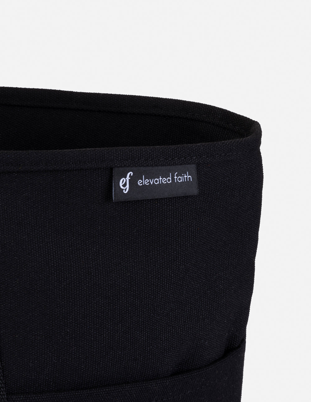 Black tote bag close-up with 'elevated faith' woven brand label on edge seam.