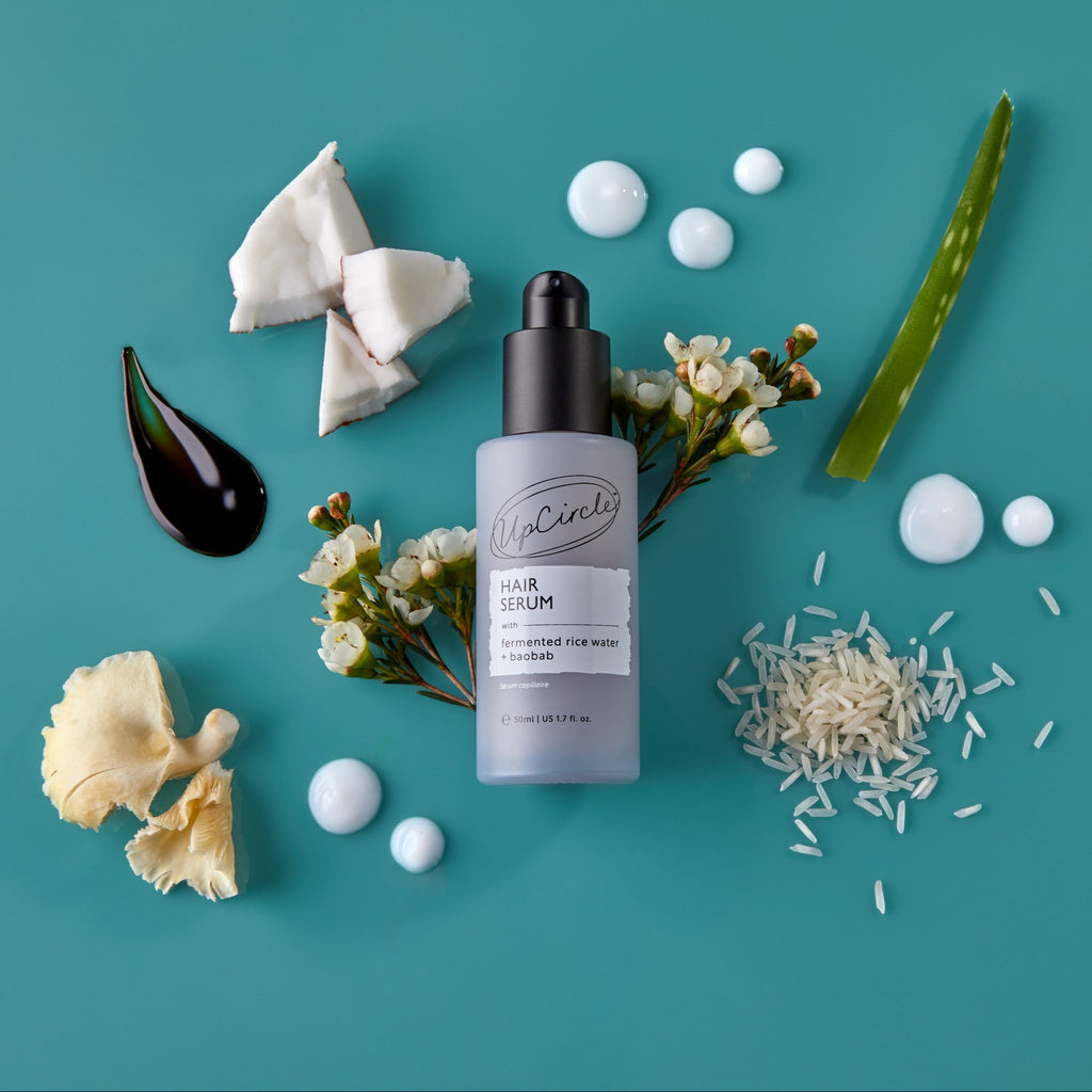 Hair serum bottle with ingredients around it, including rice grains and cream drops