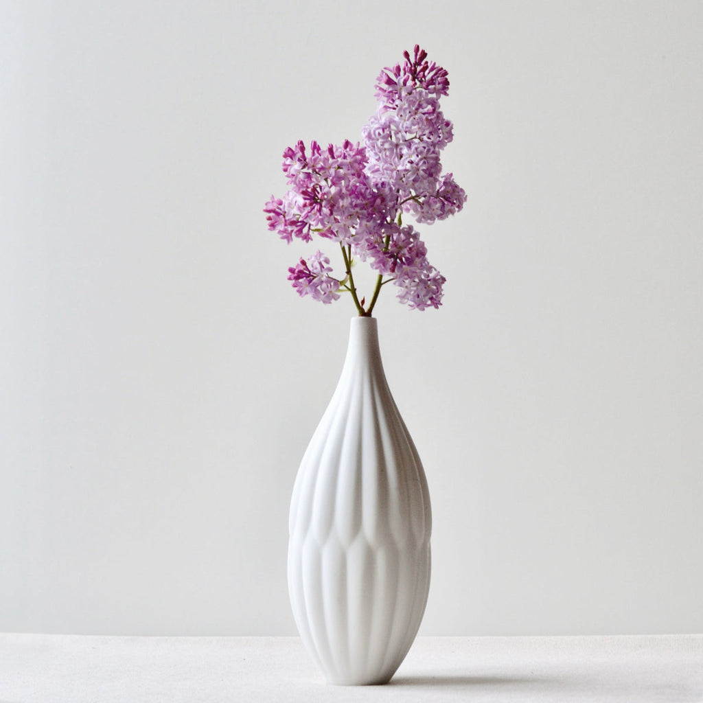White textured porcelain vase with purple lilac flowers, tall ribbed neck and body.