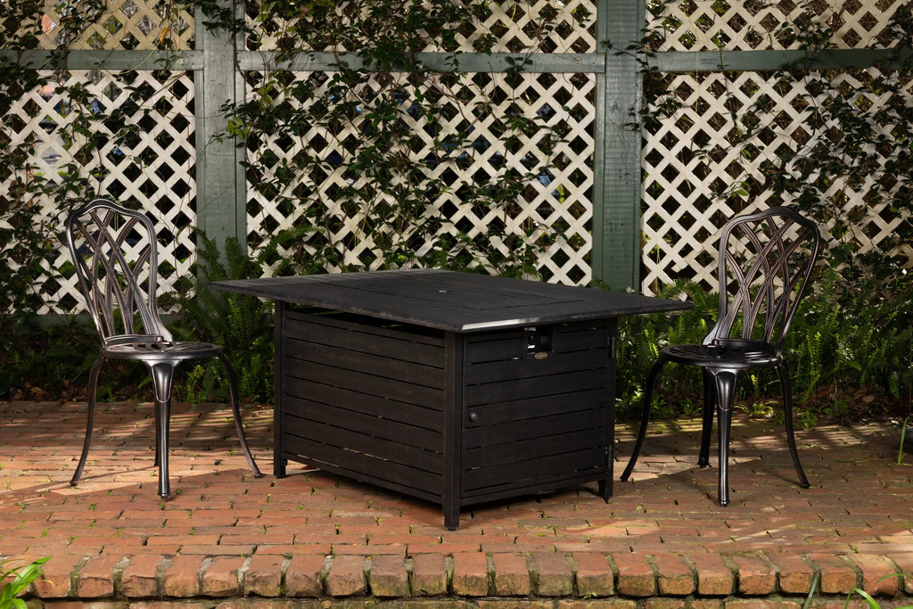 Rectangular dark gas fire pit table with open lid and two outdoor chairs nearby.
