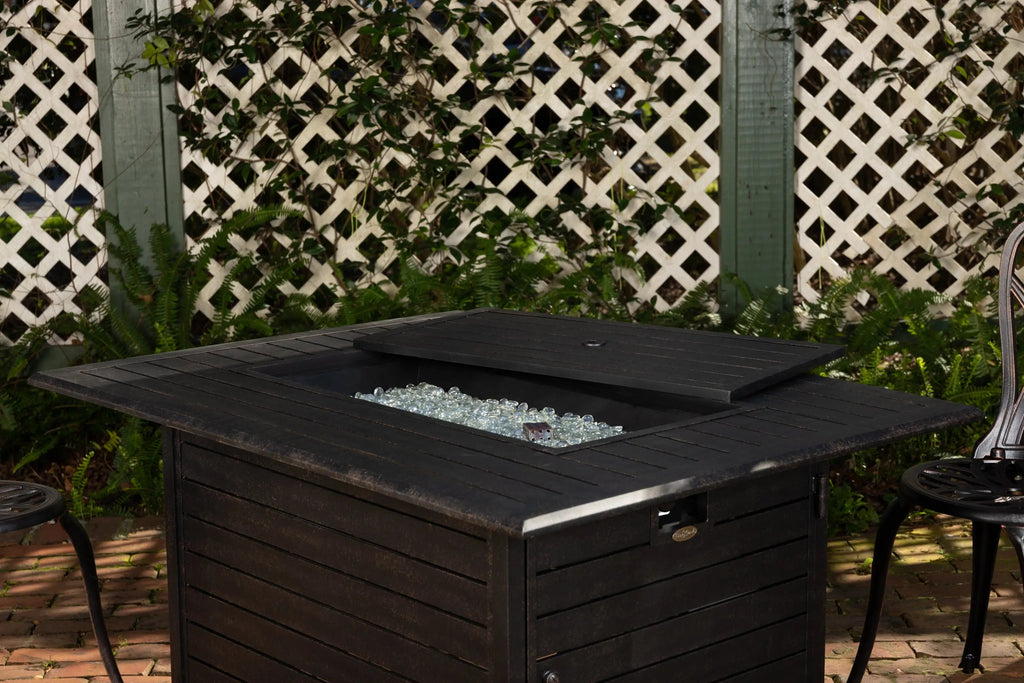 Dark rectangular gas fire pit table with open lid and glass fire media visible.