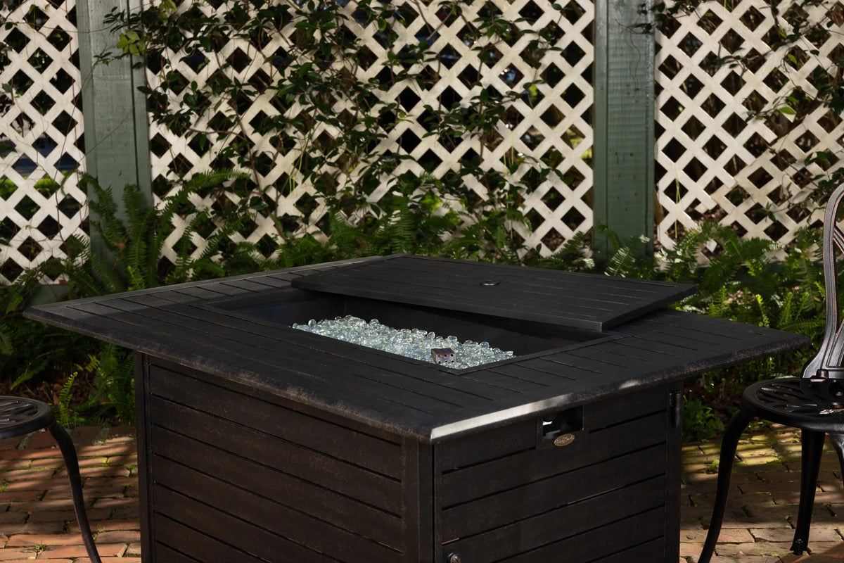 Dark rectangular gas fire pit table with open lid and glass fire media visible.