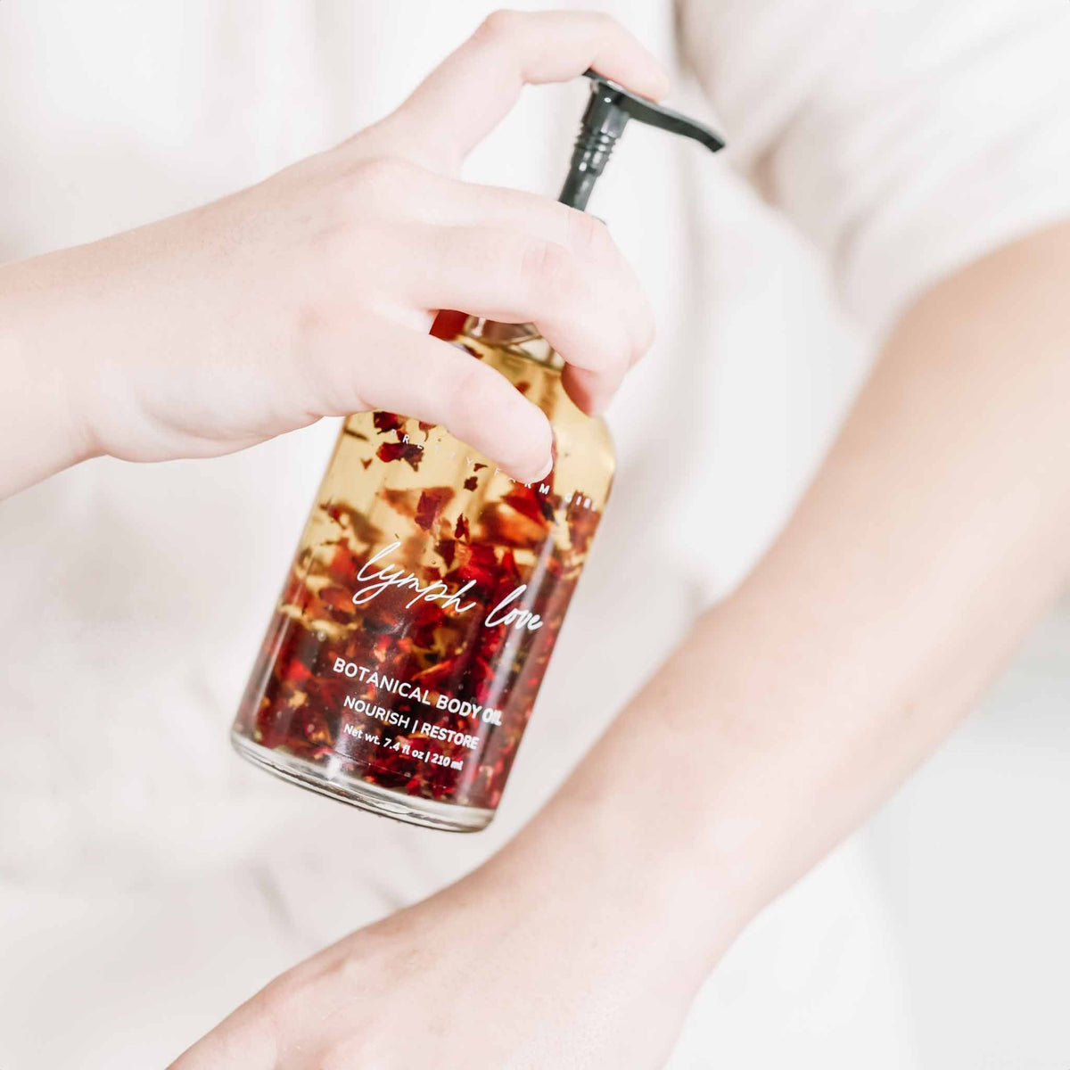 Amber botanical body oil bottle with black pump, hands holding upright.