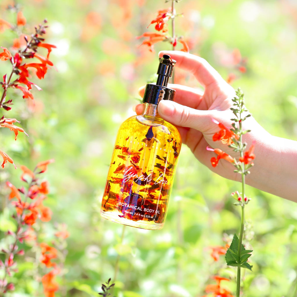 Amber botanical body oil bottle with black pump, held near orange blossoms.