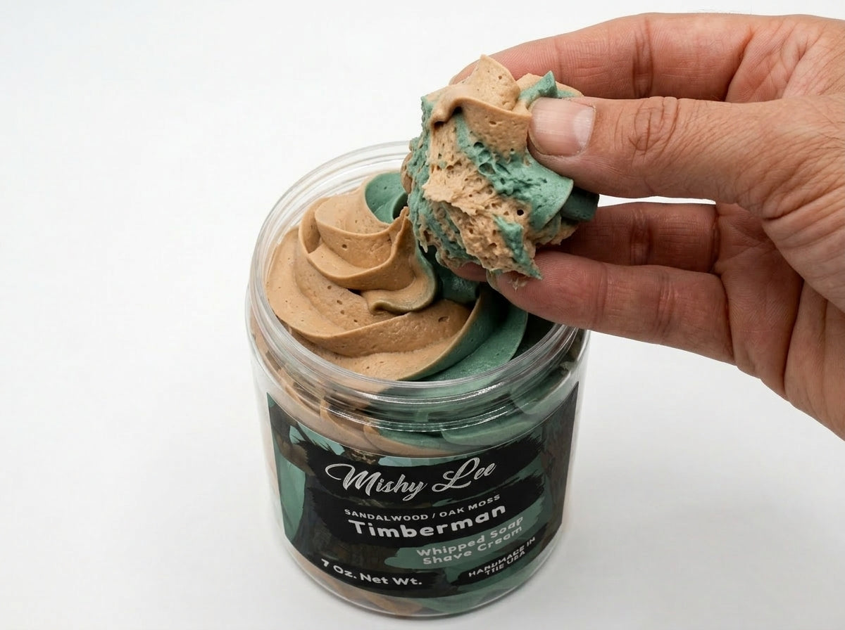 Hand holding scoop of whipped soap and shave cream from jar, showing teal and tan colors