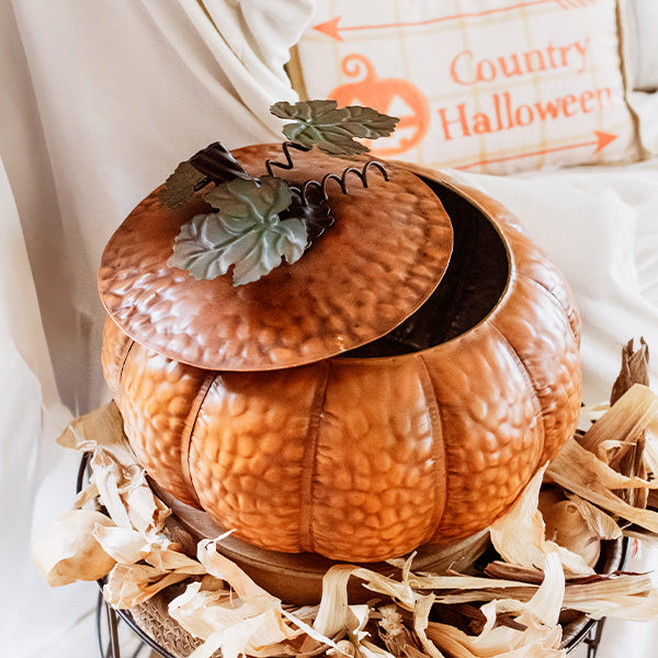 Orange metal pumpkin with open lid and green leaf topper, styled with shredded filler
