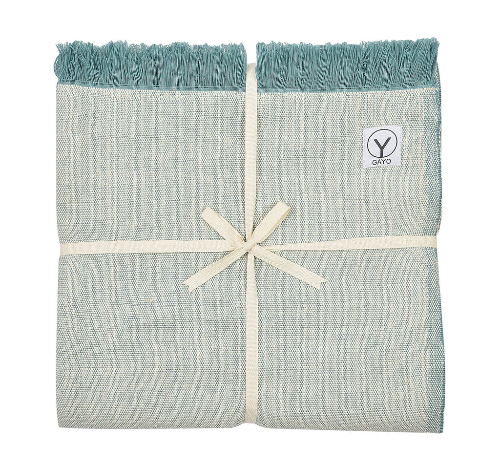 Light sage yoga blanket with teal fringe, folded bundle with logo patch and ribbon bow.