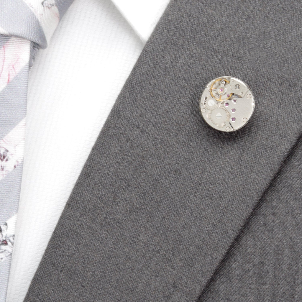 Silver watch-movement lapel pin on a grey suit jacket lapel
