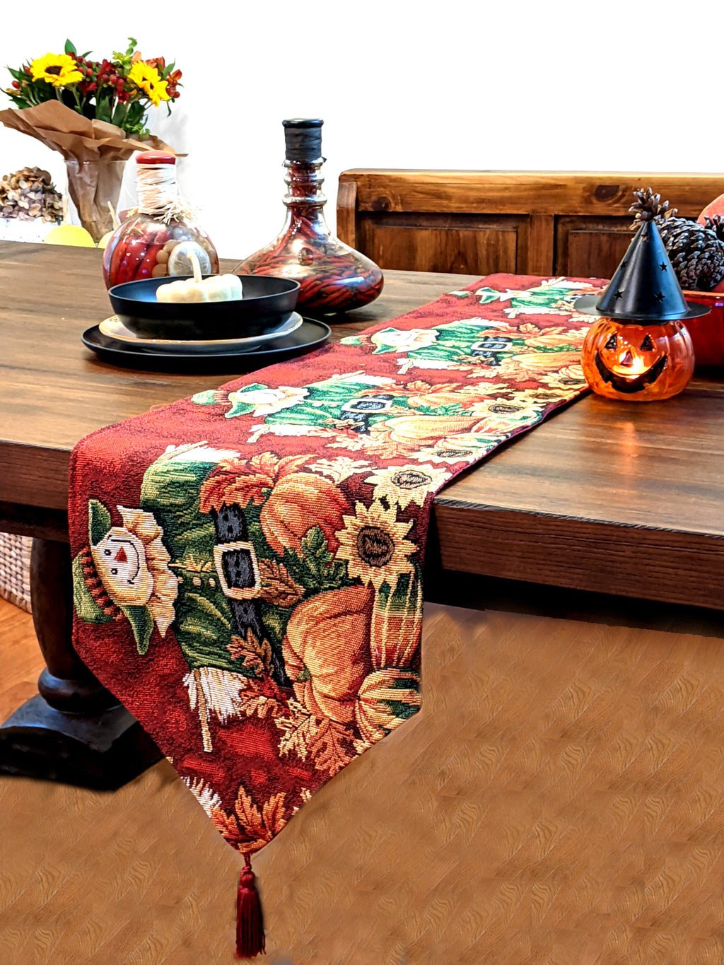 Red woven tapestry table runner with scarecrow, pumpkins, foliage, and dangling tassels.
