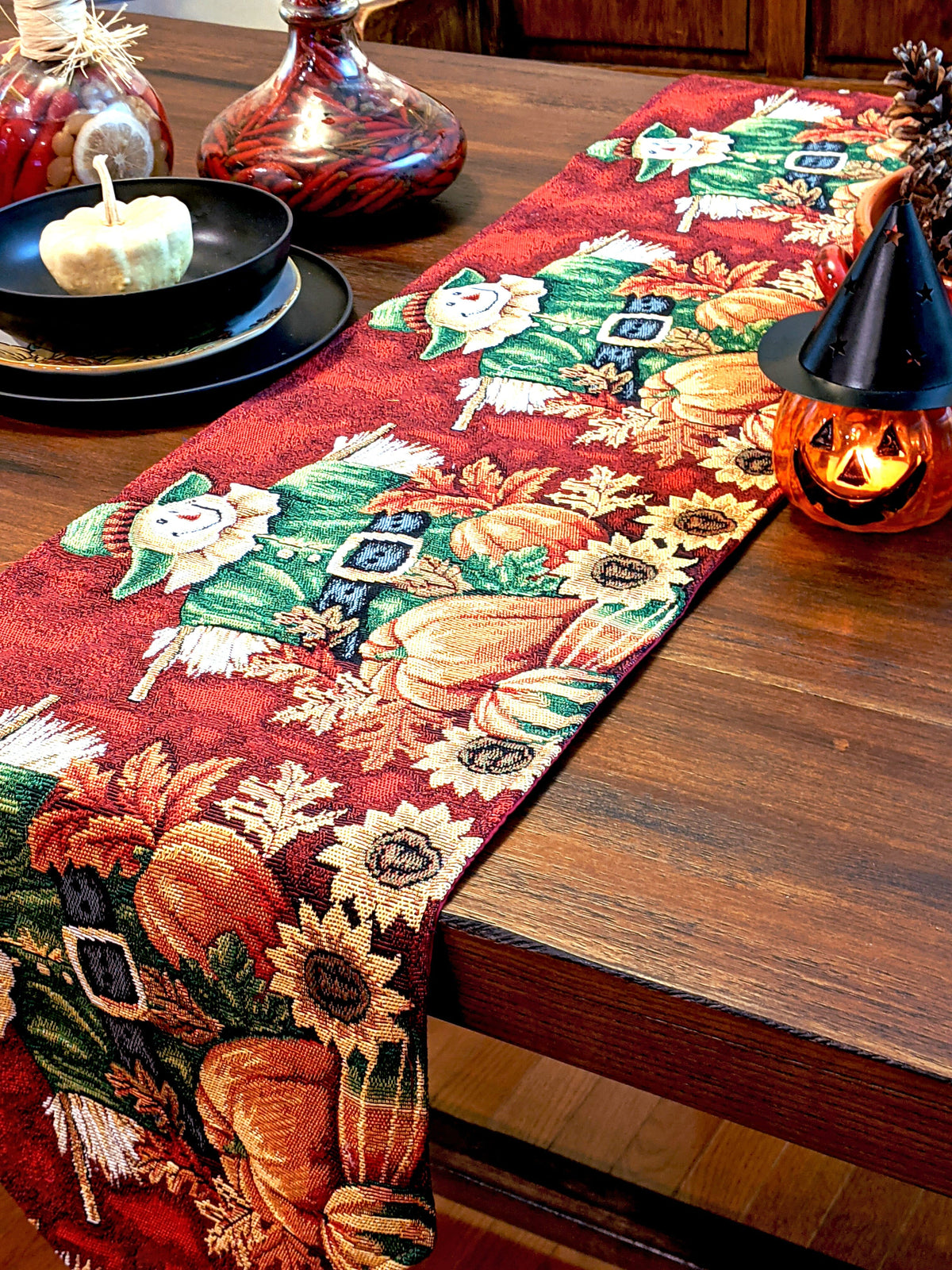 Red autumn tapestry table runner with scarecrow, pumpkins, pine sprigs, and fringed ends.