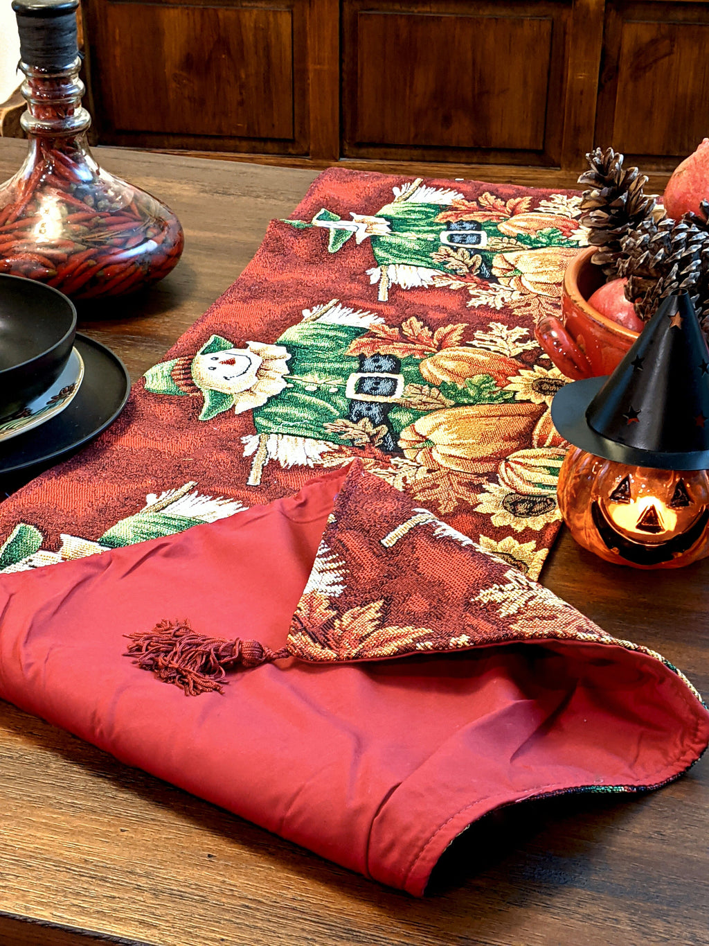 Red tapestry table runner with scarecrow, pumpkins, sunflowers, and tassel fringe.