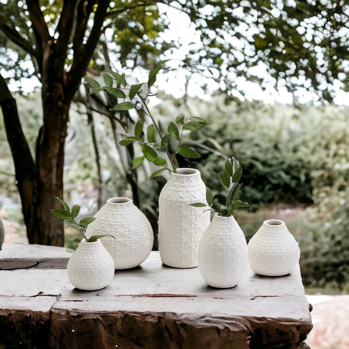 Set of white textured vases with green stems, arranged outdoors on stone