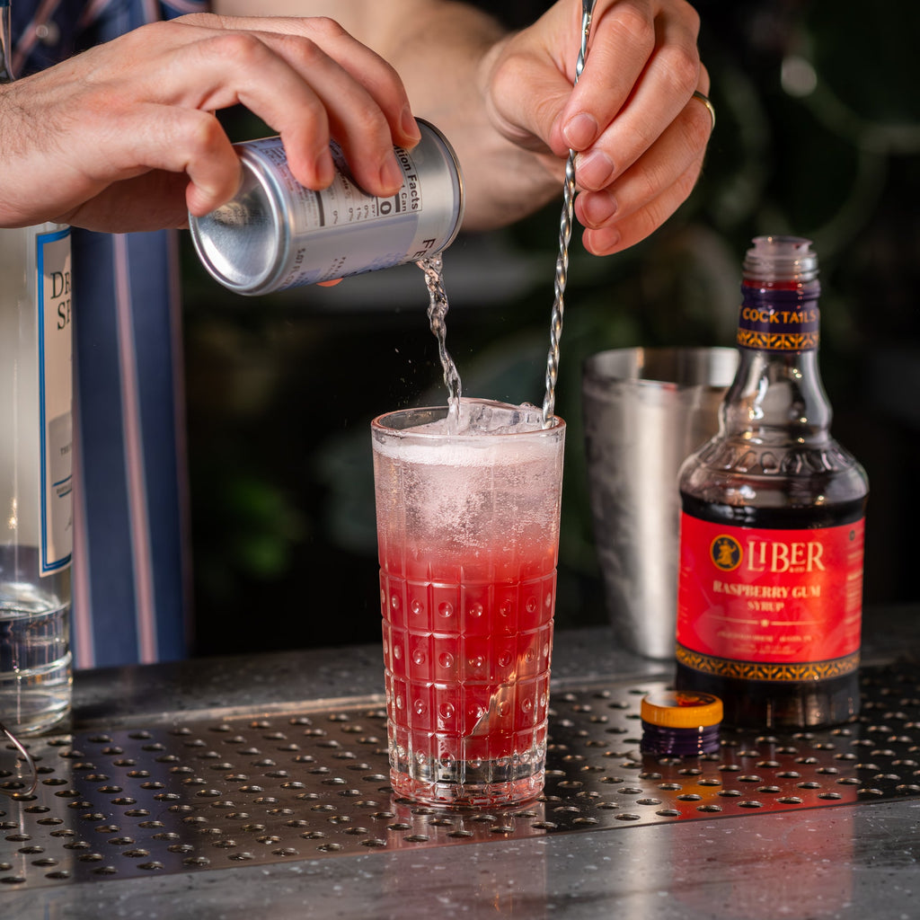 Raspberry gum syrup being poured into a red cocktail glass, Liber bottle nearby