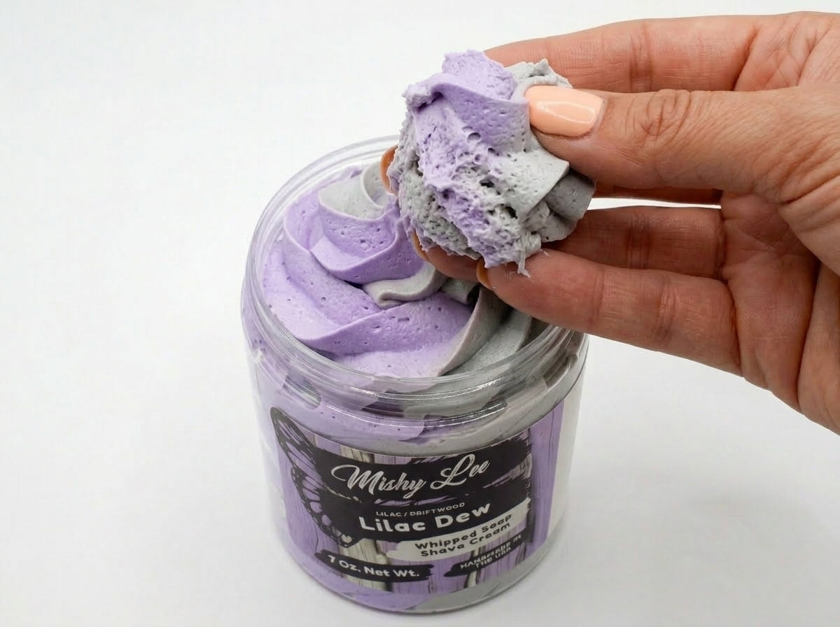 Hand holding a scoop of lilac and gray whipped soap from a clear jar