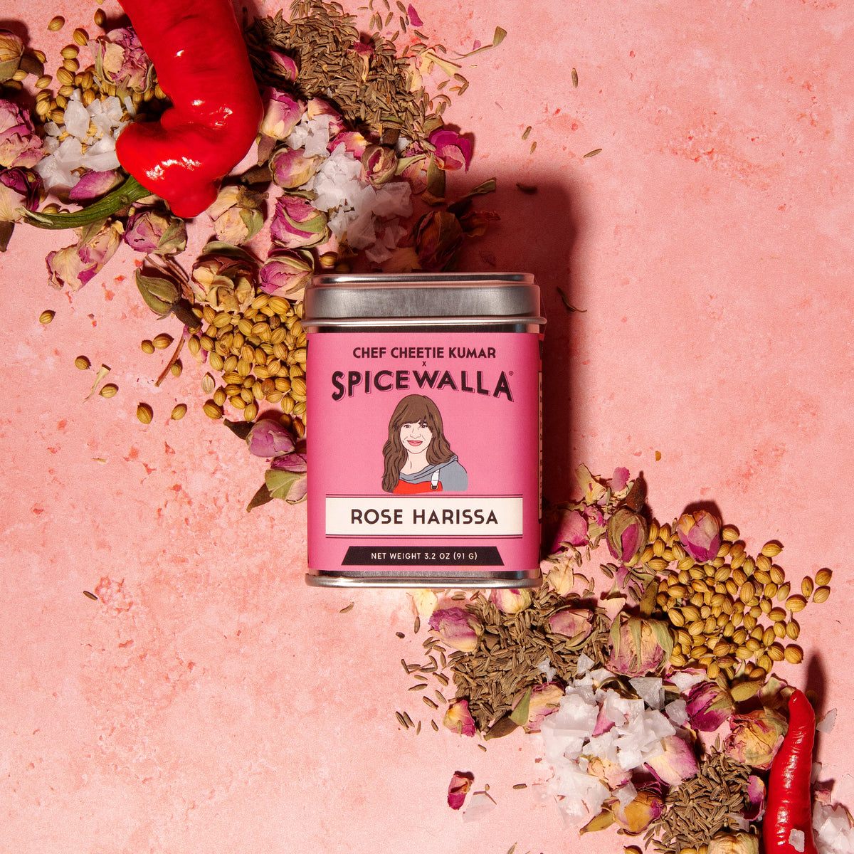 Rose harissa spice tin on pink surface with chili, seeds, and dried herbs scattered