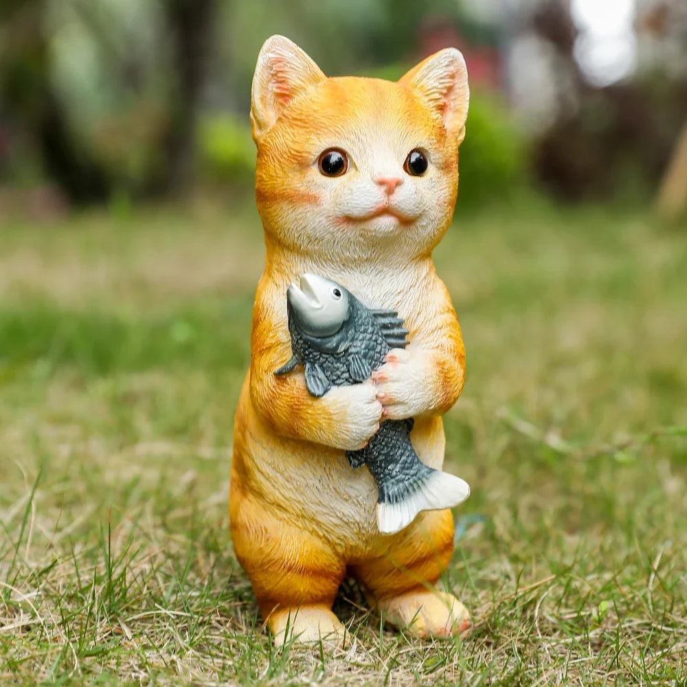 Orange cat figurine sitting on grass, holding a gray fish with both paws