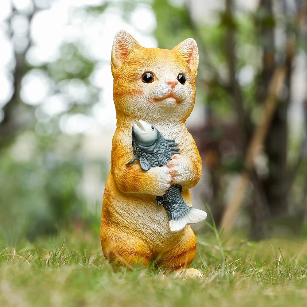 Orange cat figurine standing on grass, cradling a gray fish in its paws