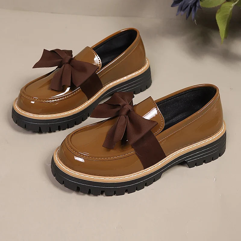 Pair of brown patent slip-on loafers with dark bow accents and thick lug platform sole.