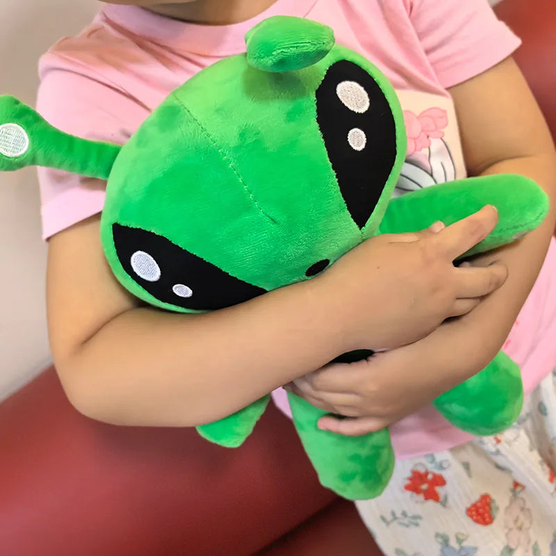 Green alien plush toy with big black eyes, held by a child’s hands on couch