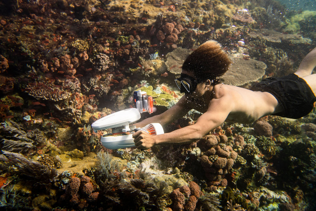 Waterproof phone case with clear front and orange seal, held underwater over coral.