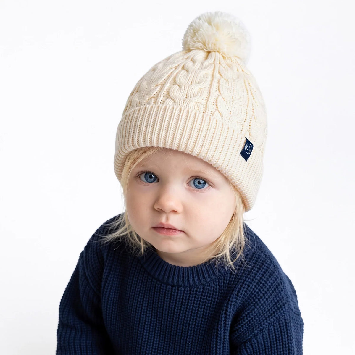 Cream cable-knit pom beanie on child model; textured cable pattern and pom top