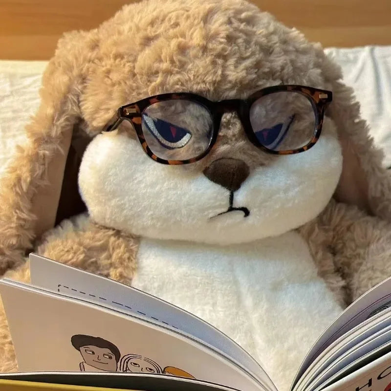 Angry long-eared rabbit plush wearing round glasses, lying beside an open book