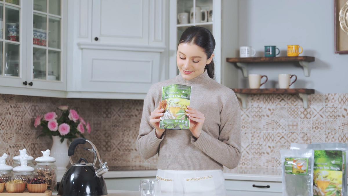 Soursop leaf tea pouch held by woman, kitchen counter with kettle