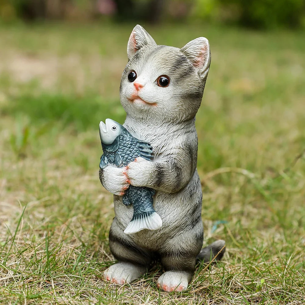 Gray cat figurine sitting on grass, holding a gray fish in its paws