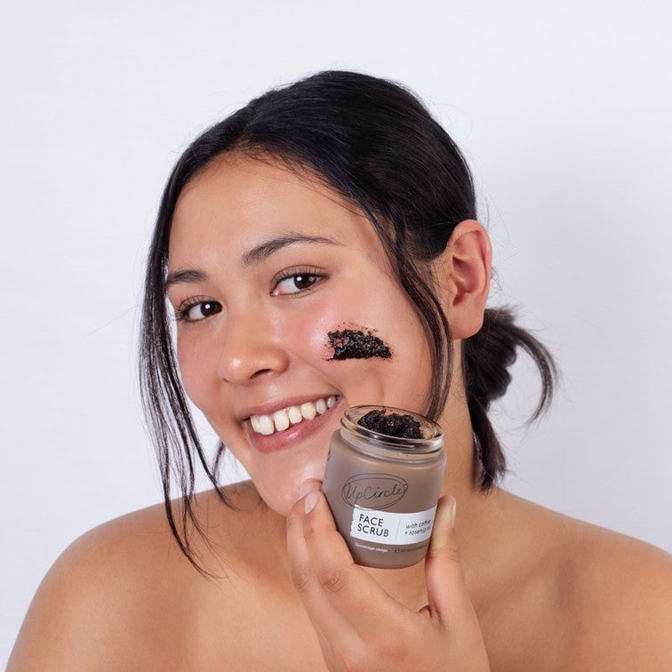 Face scrub jar with coffee and rosehip oil, held near cheek with dark scrub