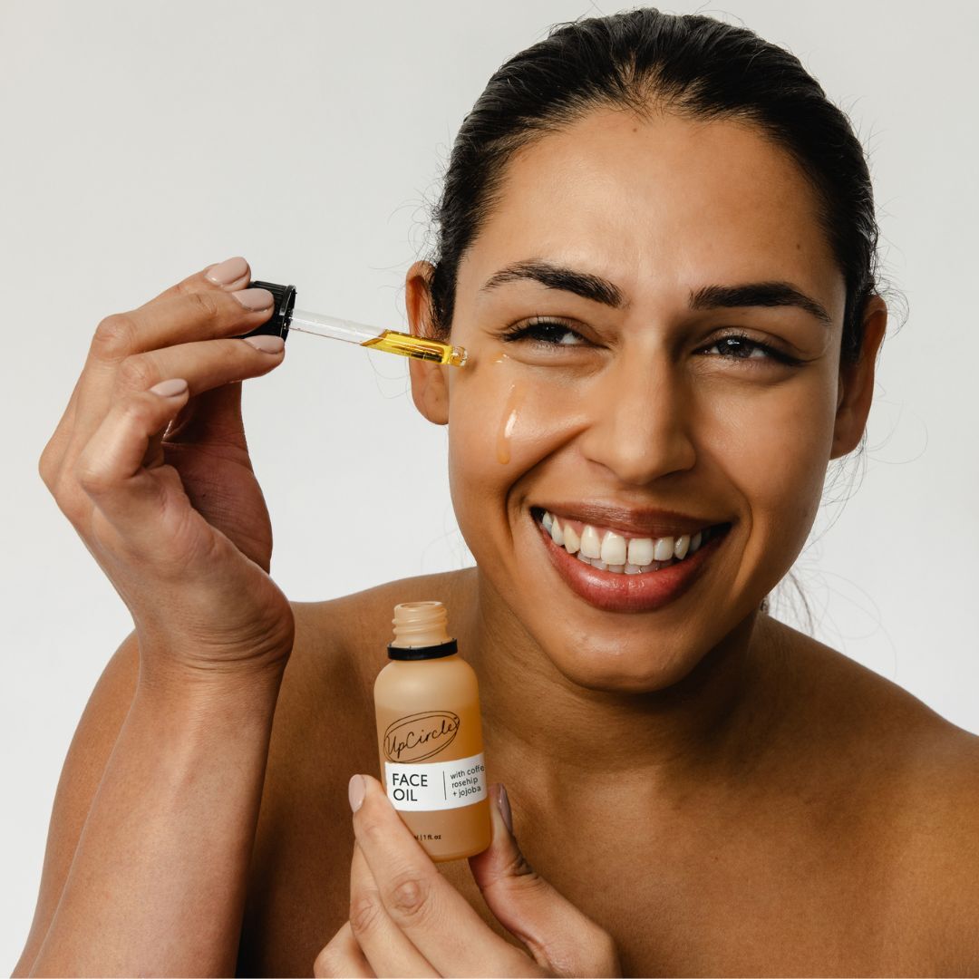 Woman applying face oil with dropper near cheek, with face oil bottle beside her