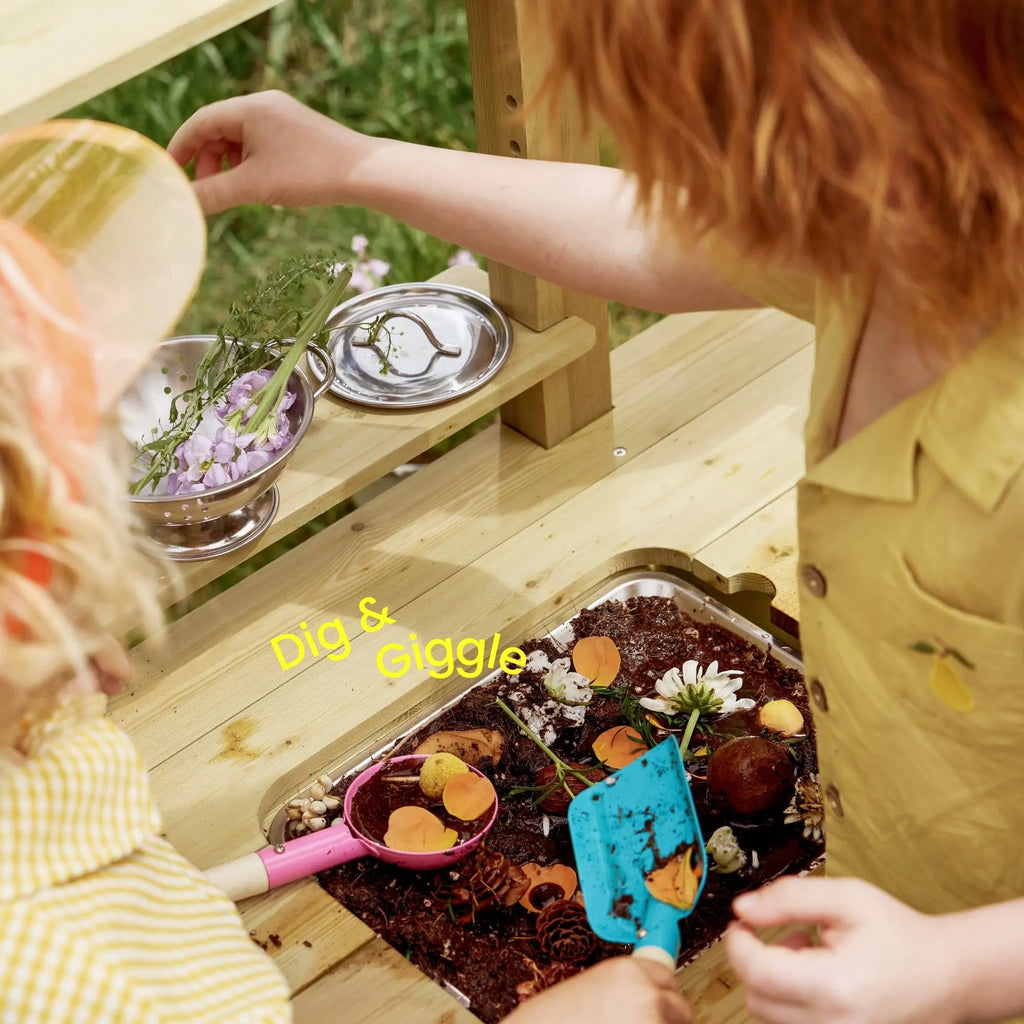 Child digs in soil tray on wooden mud kitchen playset with shovel and flower bits