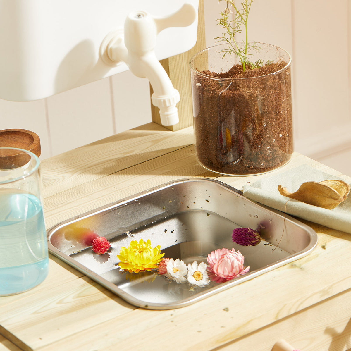 White faucet dispensing into metal tray with flowers on wooden mud kitchen playset