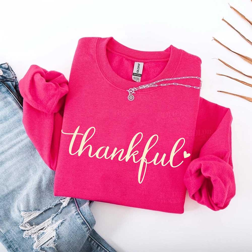 Pink crewneck sweatshirt with “thankful” script and layered chain charm graphic at collar