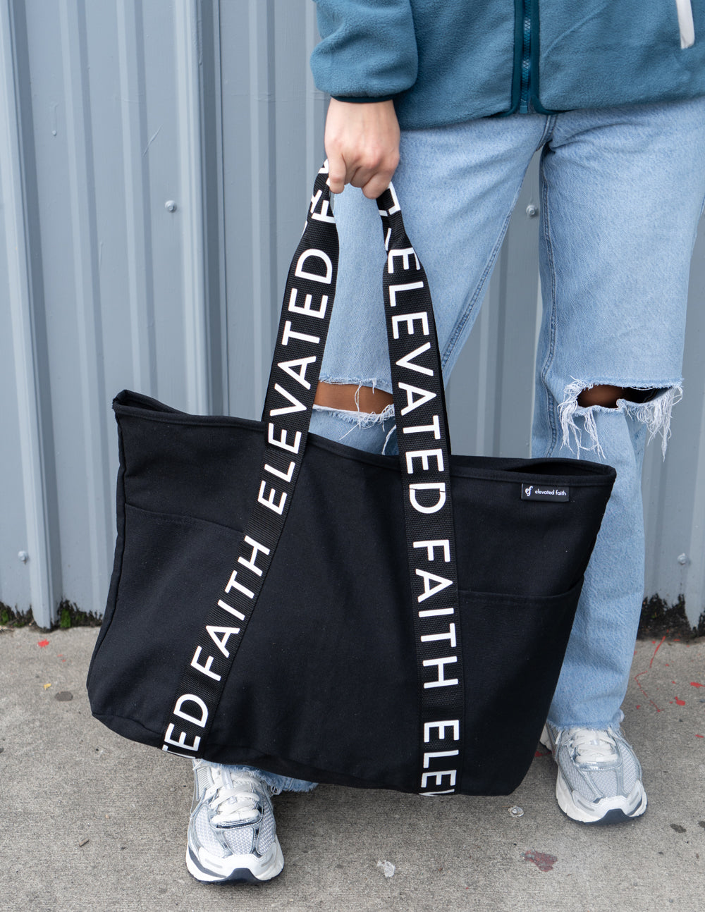 Black tote bag with long handles and vertical white 'ELEVATED FAITH' lettering on sides.
