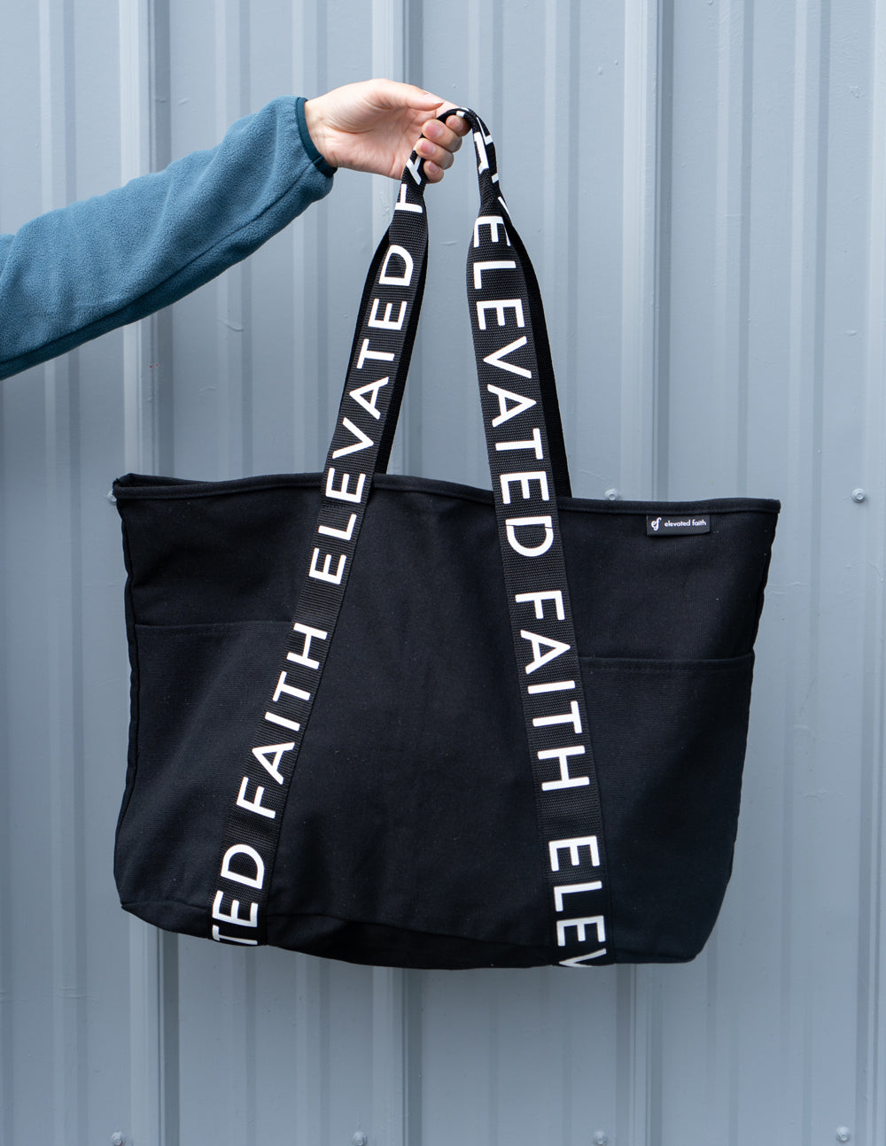 Black tote bag held by handles with vertical white 'ELEVATED FAITH' lettering.