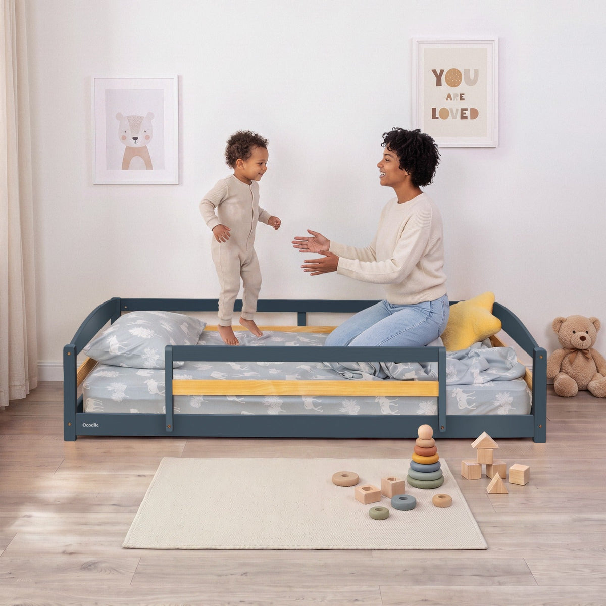Dark blue twin floor bed with safety guardrails and patterned bedding; child standing near it