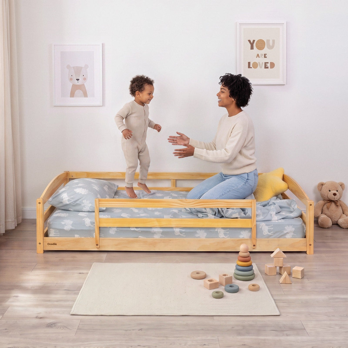 Natural wood twin floor bed with safety guardrails and patterned bedding; child climbing in