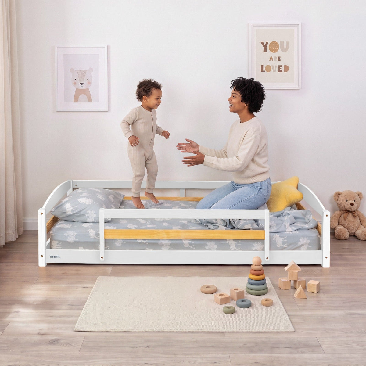 White twin floor bed with safety guardrails and patterned bedding; child reaching toward the bed
