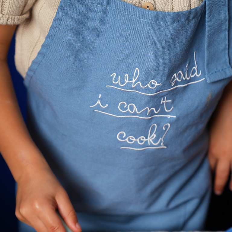 Blue cooking apron close-up with embroidered white script lettering on bib front