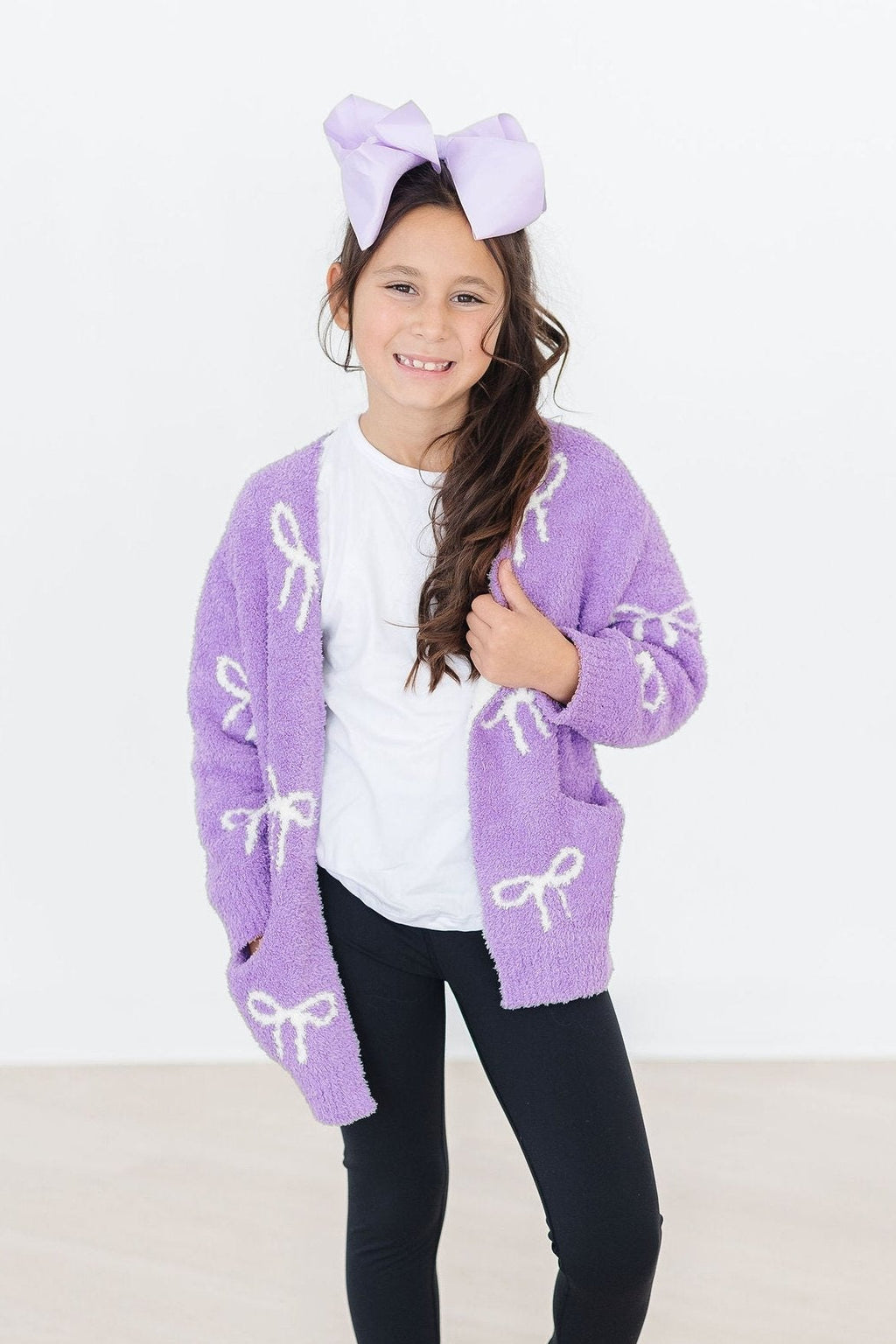 Girls purple plush cardigan with bow pattern, open front, worn over white top.