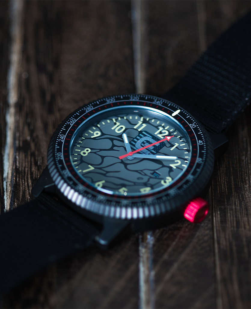 Round camo wristwatch with dark bezel, red accent crown, and black fabric strap