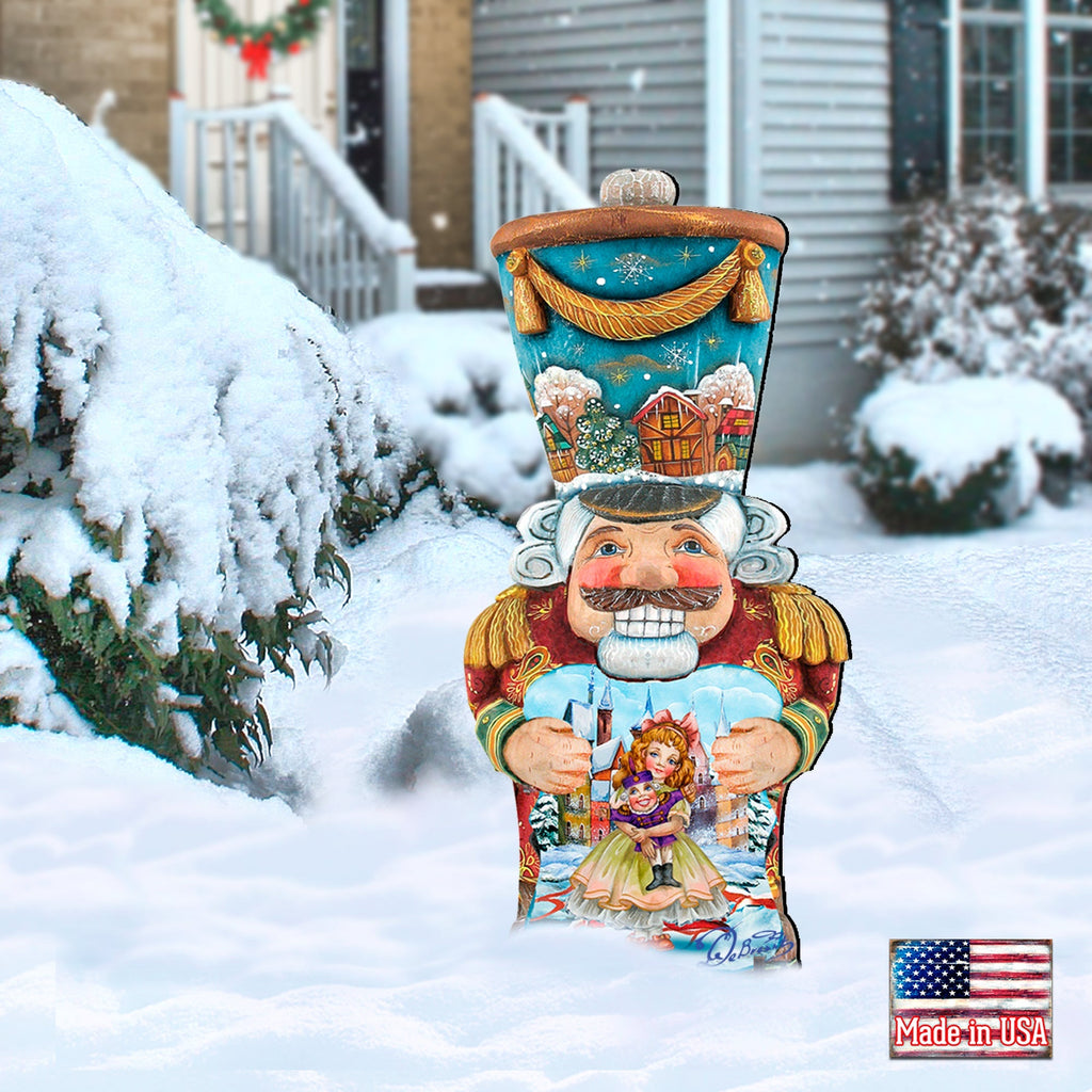 Painted nutcracker yard decor with blue top, red coat, holding doll, standing in snow